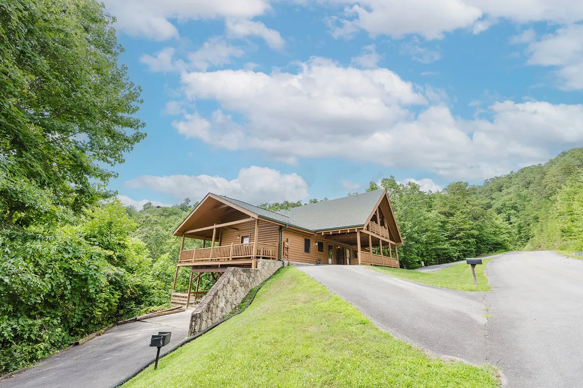 The exterior of a welcoming wooden cabin nestled among verdant greenery beneath a soft, cloudy sky. The inviting porch and surrounding landscape hint at a retreat perfectly situated for family relaxation after a day of exploration.