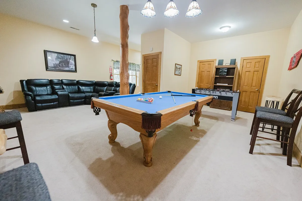 This vibrant game room scene features a pool table at the center, a plush leather sofa, bar stools tucked to the side, and a foosball table in the background. The room is well-lit, with plenty of space for groups to gather, play, and socialize. The arrangement of different gaming options, comfortable seating, and open layout highlights the area as the perfect entertainment retreat.