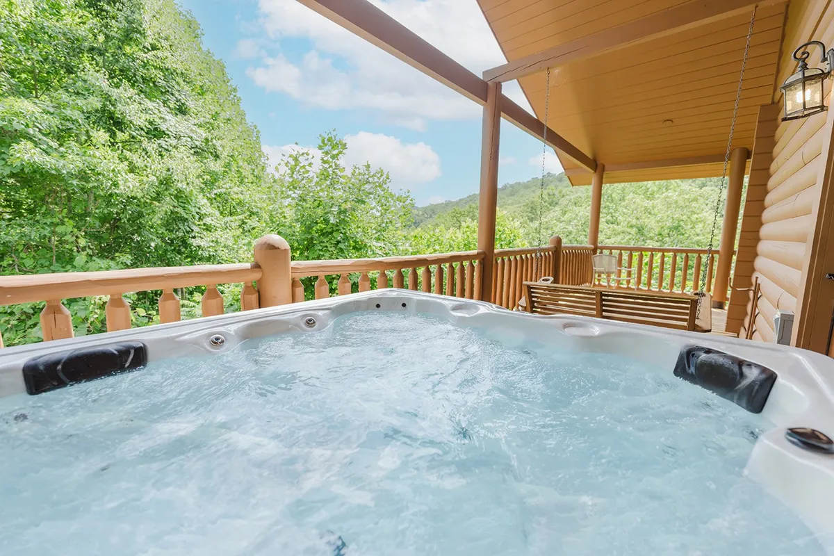 This image features a bubbling hot tub set on a wooden deck, flanked by a swing bench and wooden railing. The backdrop is a lush canopy of green trees, creating a private oasis for relaxation. Both the swing and hot tub are prominent, with sunlight filtering through the trees, enhancing the tranquil mood.