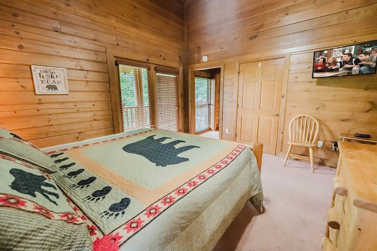 A cozy bedroom featuring a bed with a bear-themed quilt, large windows with blinds, rustic wooden furniture, and a TV mounted on the wall. Natural light spills into the space, emphasizing both comfort and a connection to nature.