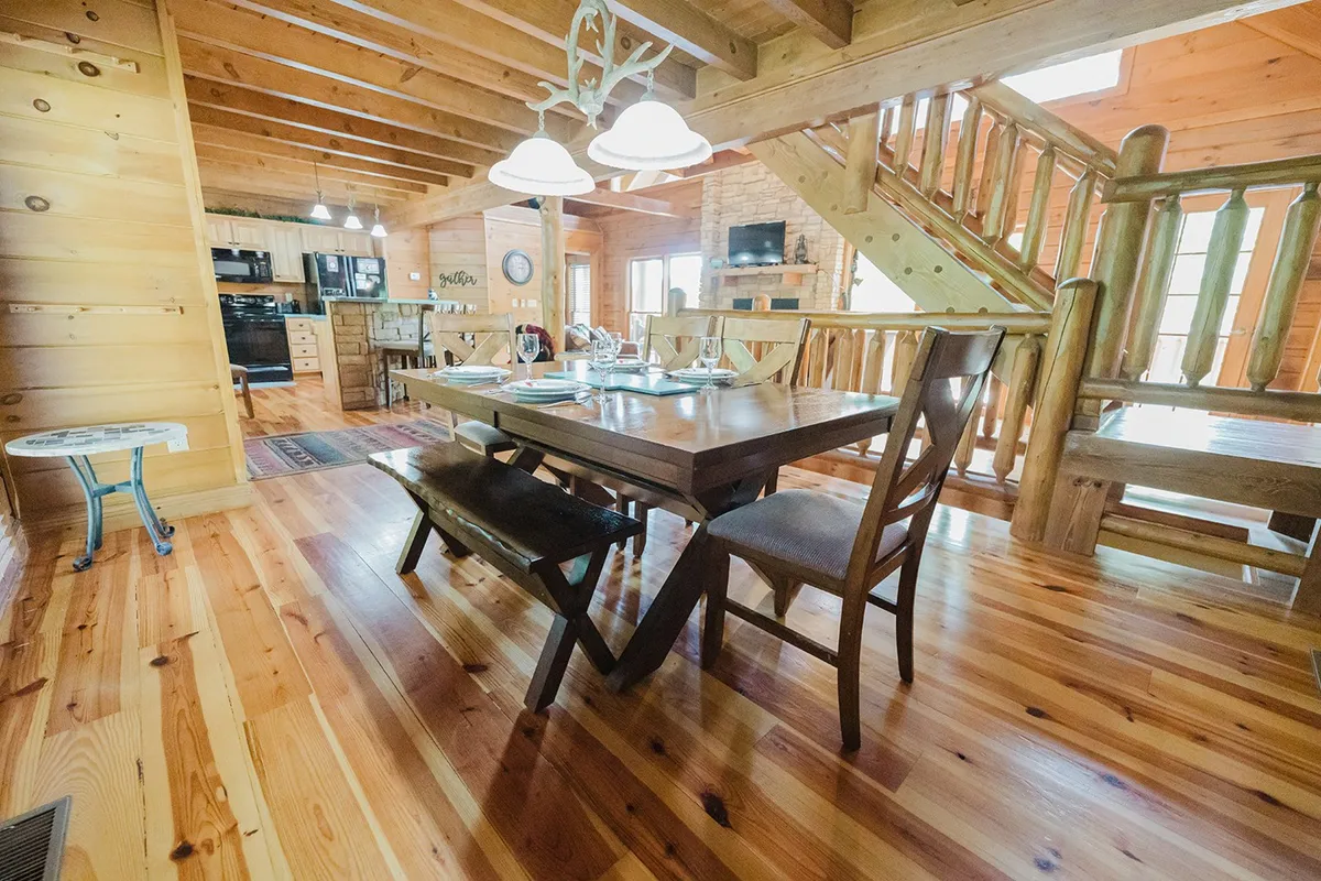 A warm dining area centered around a large wooden table, with surrounding wooden elements, cozy lighting from chandeliers, a visible fireplace, and an adjacent staircase. The setting is ideal for shared meals, games, or holiday celebrations.