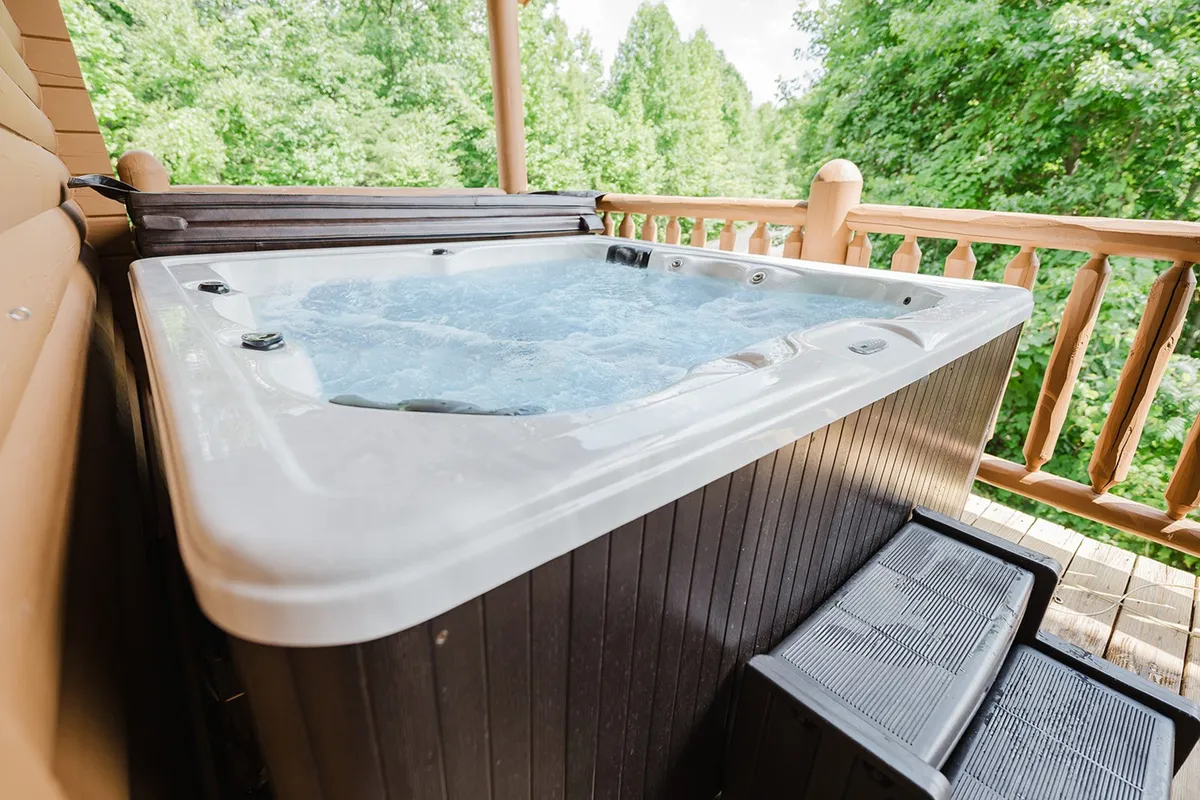 An outdoor hot tub sits on a spacious wooden deck, bordered by a rustic railing and surrounded by trees. The serene setting suggests moments of relaxation under the stars after busy days of family fun and exploration.