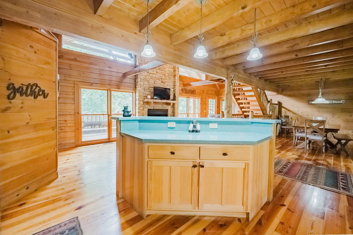 A spacious open-concept area connecting a kitchen island, a dining table, and a living area with a stone fireplace. Large windows bring in natural light, and the kitchen is fully equipped with modern appliances and ample counter space, seamlessly integrating dining and cooking spaces for group interaction.