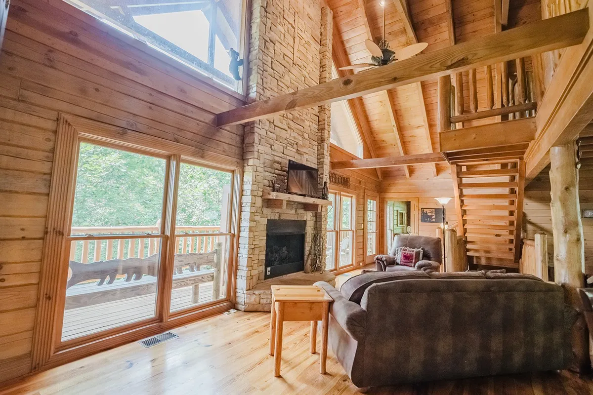 A welcoming living area highlights a stone fireplace surrounded by comfortable seating and large windows that reveal scenic views. The wooden staircase and natural accents add a rustic charm, perfect for gatherings and relaxation after exploring the outdoors.