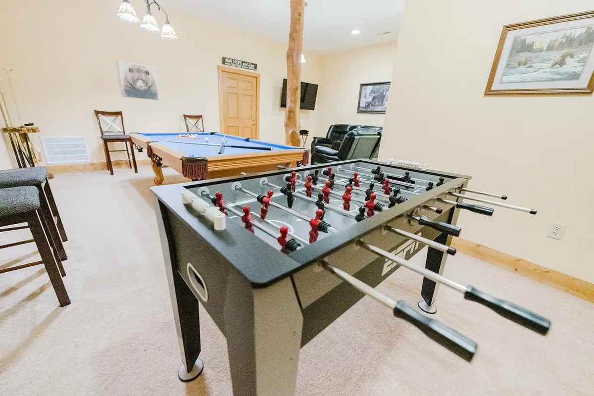 A playful snapshot of the entertainment area featuring a foosball table and pool table side-by-side, with comfortable recliner chairs and a television nearby. The setup encourages both active play and relaxed spectating, allowing for varied entertainment options in a lively, cozy environment.