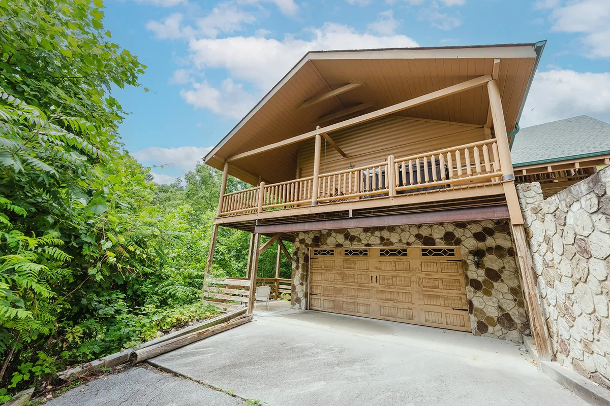 This exterior shot features a spacious balcony overlooking a lush green landscape, with a garage and stone wall visible. The surrounding forested area and open sky evoke a strong sense of connection to nature and the outdoors.