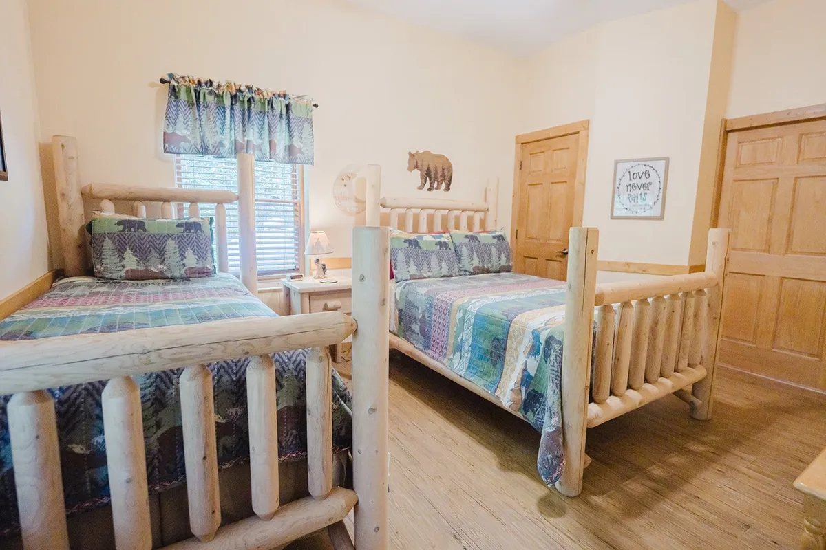 A lower-level bedroom with log beds adorned with colorful quilted bedding, wooden furniture, a bear decoration, and a window with curtains letting in daylight. The room is warm, inviting, and ideal for families or groups of friends.