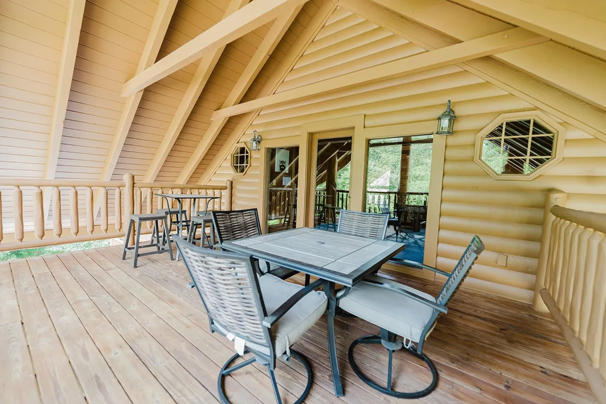This image showcases a spacious outdoor dining area on a wooden deck, complete with a sturdy table, wooden railing, and large windows looking back into the cabin interior. The area is enveloped by lush greenery, evoking a sense of seclusion and peace, perfect for alfresco meals and gatherings as described in the section.