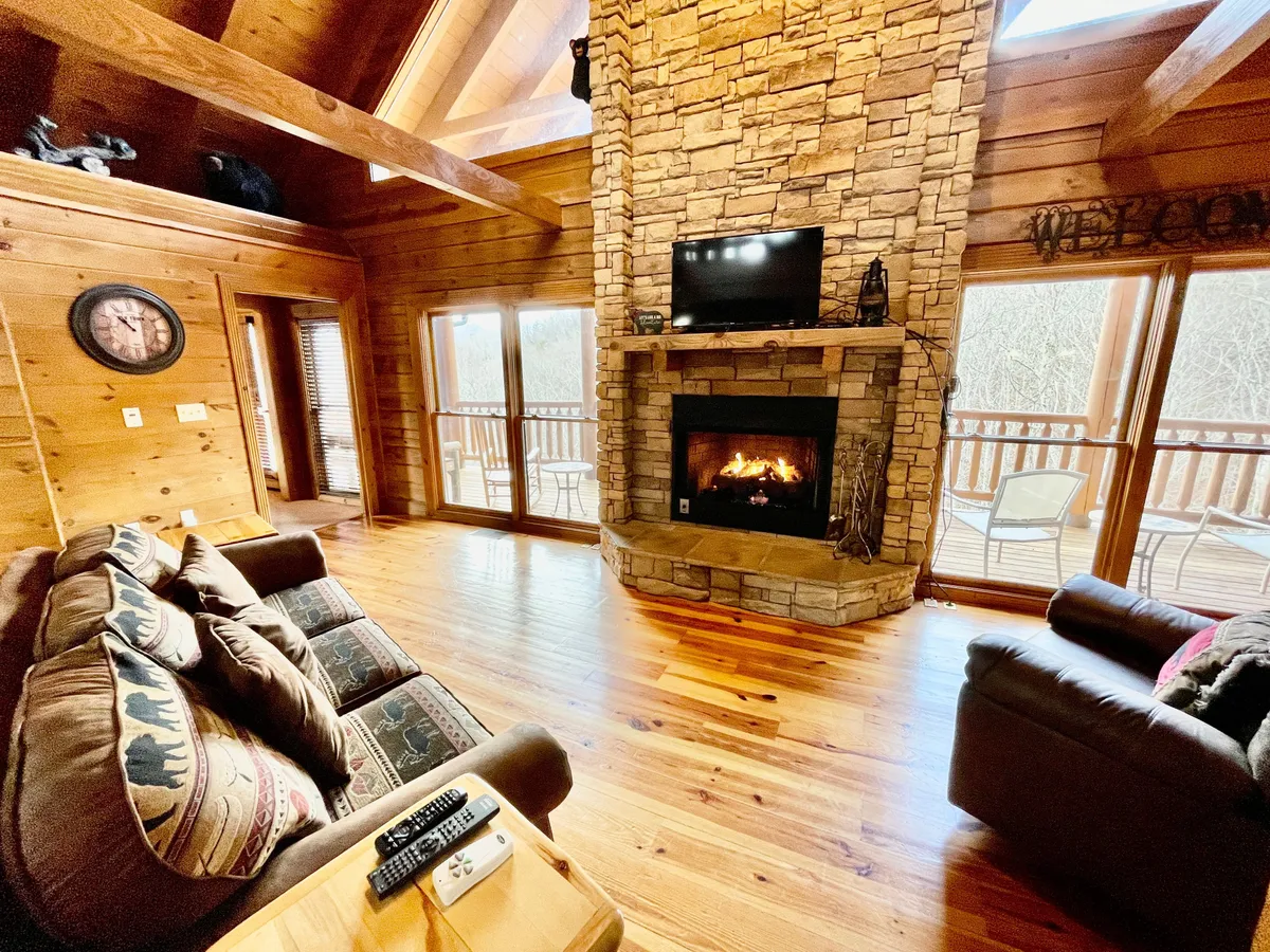 A warm, inviting living room featuring a stunning stone fireplace, large windows with scenic views, exposed wooden beams, and plush seating arranged for conversation and relaxation. The TV is positioned near the fireplace, making this space ideal for movie nights or cozy gatherings.